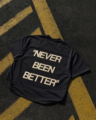 A black short-sleeve t-shirt with the phrase "NEVER BEEN BETTER" printed in large beige block letters on the back, laid flat on asphalt over yellow road markings.