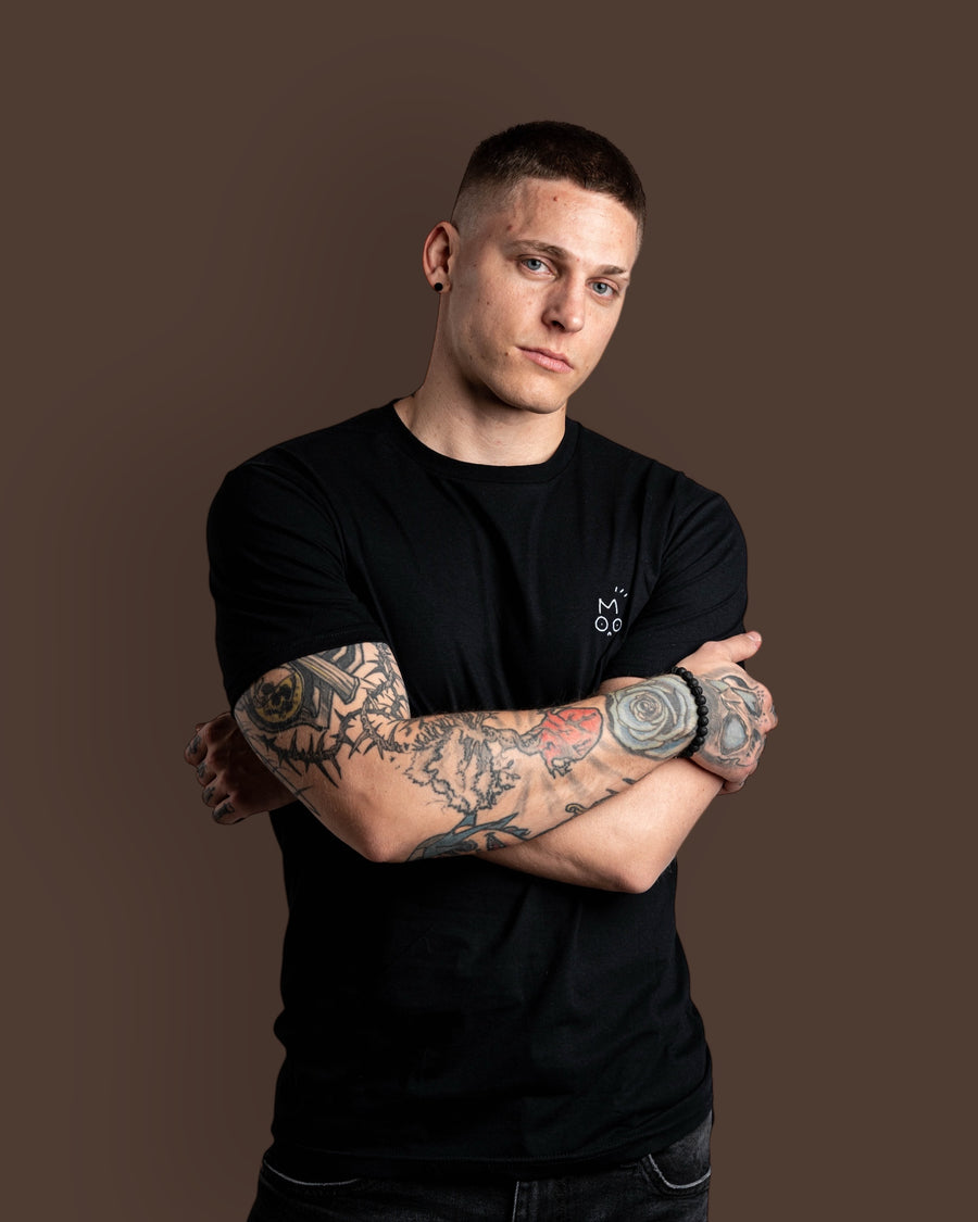 Man with tattoos wearing a black streetwear OUT OF MOOD SIMPL t-shirt against a brown background