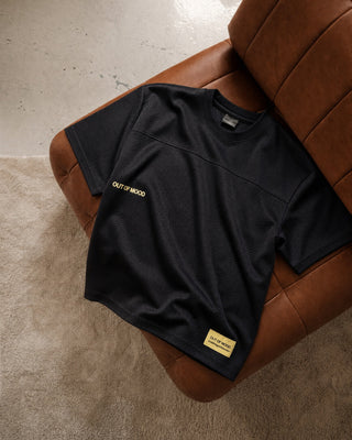 A black short-sleeve athletic t-shirt with gold text reading 'OUT OF THE MOOD' and a small gold brand patch, laid flat on a brown leather armchair over a beige carpet.
