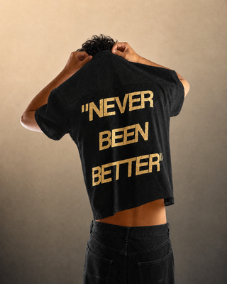 A person shown from behind wearing a black cropped T-shirt with the phrase "NEVER BEEN BETTER" printed in large gold letters on the back, paired with dark jeans against a plain beige studio background.