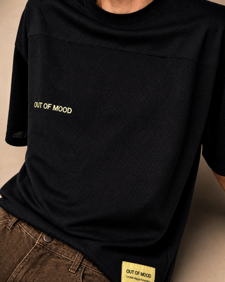 A close-up of a person wearing a black textured short-sleeve t-shirt with 'OUT OF MOOD' printed in yellow on the chest and a matching yellow patch on the hem, paired with brown jeans.
