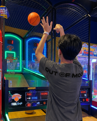 A man wearing a dark gray t-shirt with 'OUT OF MOOD' printed on the back, standing in an arcade and shooting a basketball toward a neon-lit basketball machine.