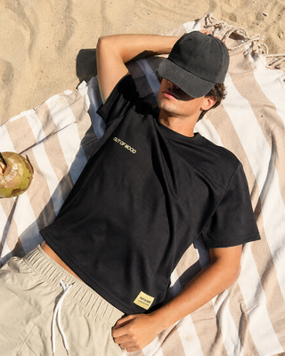 A man lying on a striped beach towel on sand, wearing a black t-shirt with gold 'OUT OF MOOD' text on the chest, a black baseball cap, and beige shorts.