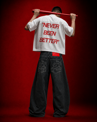 A model from behind wearing a white cropped t-shirt with 'NEVER BEEN BETTER' printed in red on the back, paired with dark wide-leg jeans, holding a red baton against a dark red background.