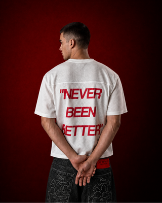 A man stands with his back to the camera wearing a white t-shirt with 'NEVER BEEN BETTER' printed in red on the back, paired with dark patterned shorts against a dark red studio background.